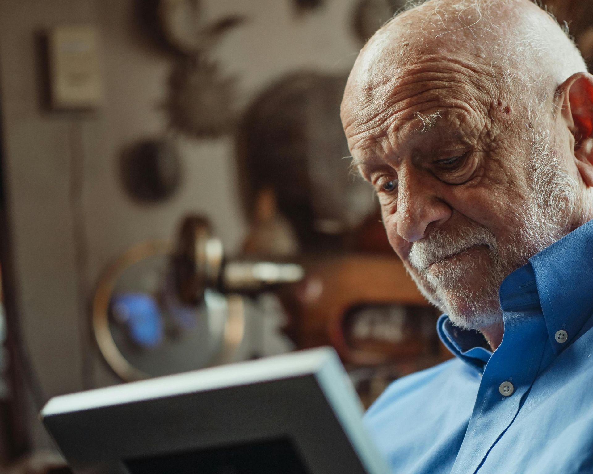 A senior man in a blue shirt looks thoughtfully at a framed photo indoors.