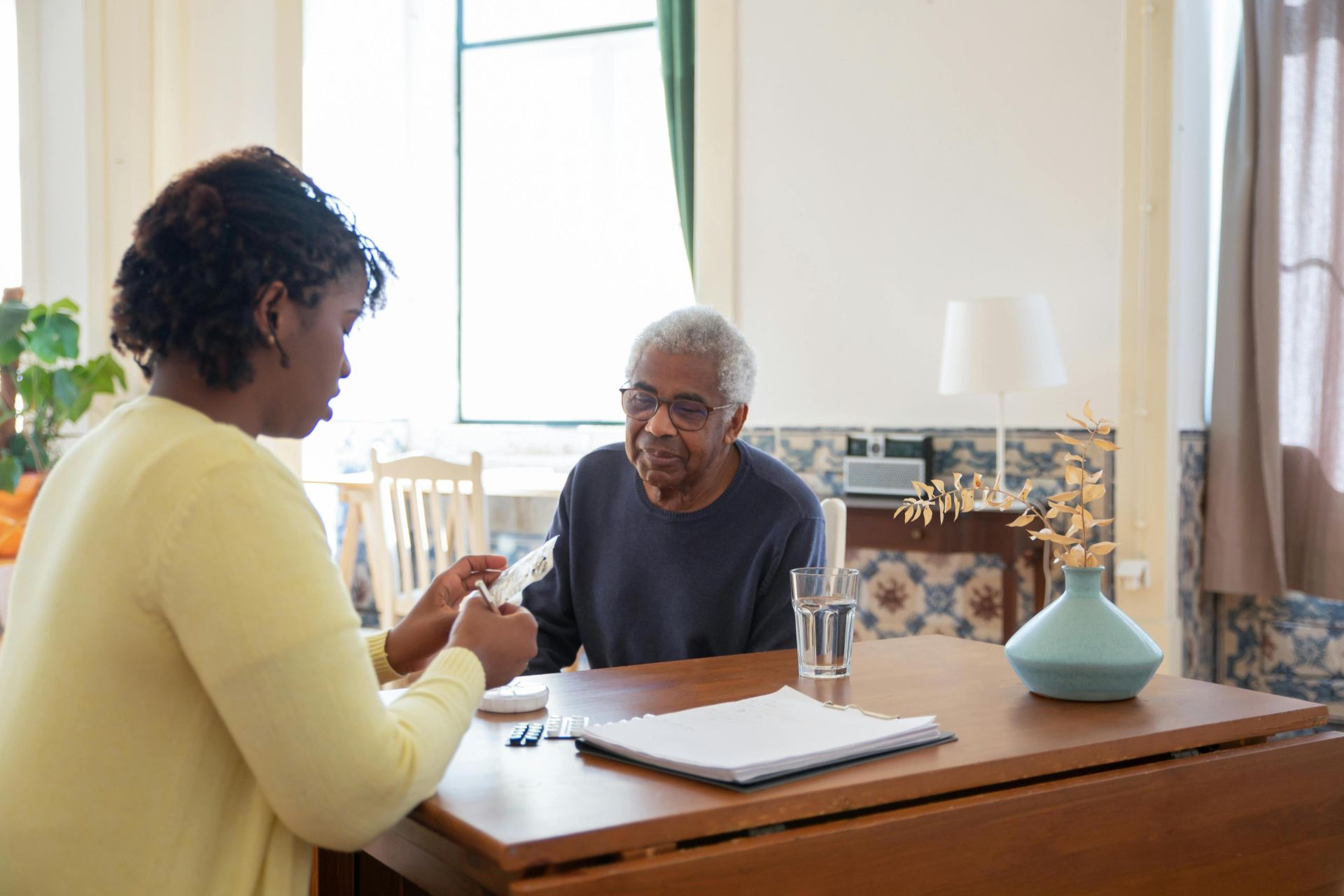 A caregiver assists an elderly man with medication in a warm, sunlit living room.