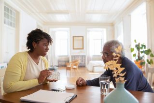 An elderly man sits with a caregiver discussing medication at a table in a bright room.