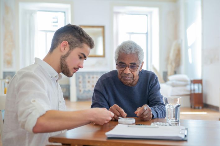 Young man assisting elderly man with medication in a bright living room setting.