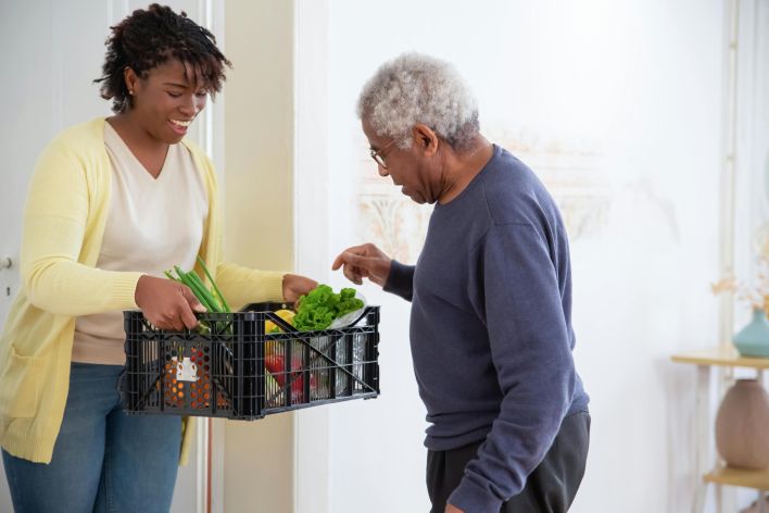 A young adult woman helps a senior man by carrying a crate of fresh vegetables indoors, symbolizing care and support.